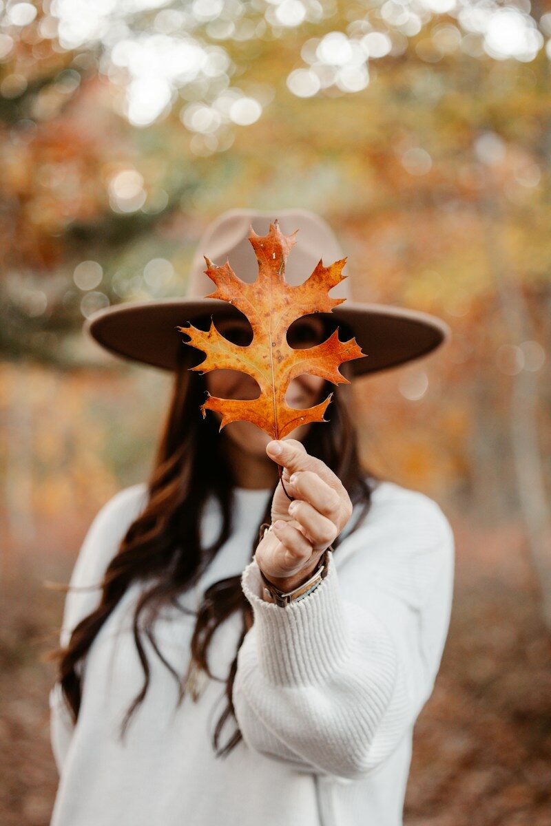 A person in a hat holding an orange oak leaf over their face